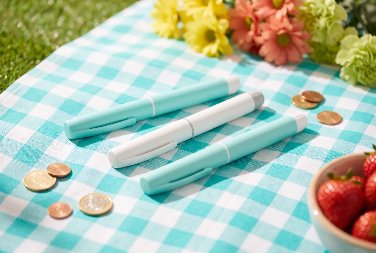 Plain injection pens in pastel colors with coins on a turquoise picnic blanket